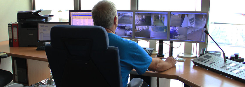 Person sitzt an einem Schreibtisch vor mehreren Monitoren mit Überwachungskamera-Bildern